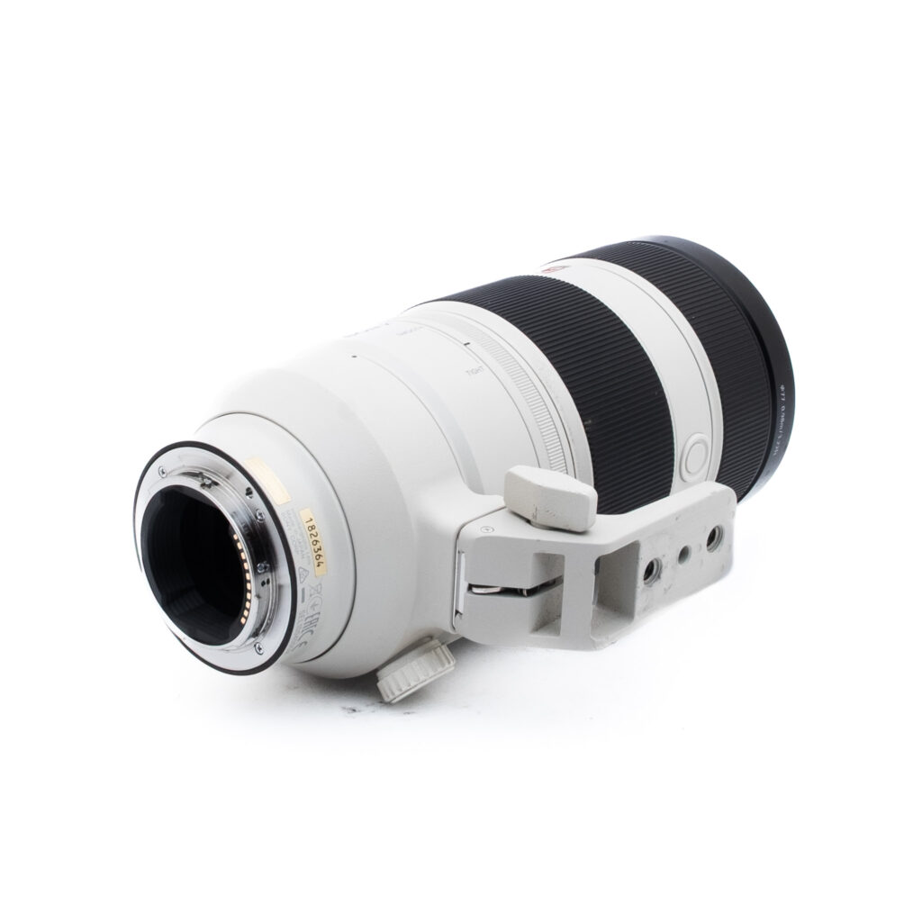 Sony 100-400 GM-1 Valkoinen telekameran objektiivi, jossa on musta tarkennusrengas, jalustan kaulus ja kiinnitysteline, kuvattuna kyljellään valkoista taustaa vasten.