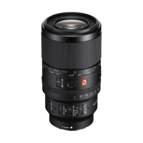 Sony FE 100mm f/2.8 Macro GM -objektiivi on musta kameraobjektiivi, jossa on kuvioidut tarkennus- ja zoomausrenkaat, aukkomerkinnät, säätökytkimet ja oranssi G-logo, joka on kuvattu pystyasennossa valkoisella pohjalla.