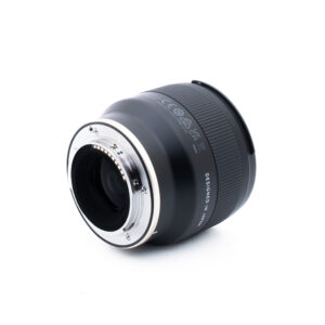 Tamron 35mm f28 Di III OSD M12 Sony-1 Lähikuva kameran objektiivista, jossa näkyy sen metallinen objektiivin kiinnitys ja elektroniset koskettimet valkoisella pohjalla. Objektiivin tynnyri on musta, ja siinä on näkyviä merkintöjä ja tekstiä.