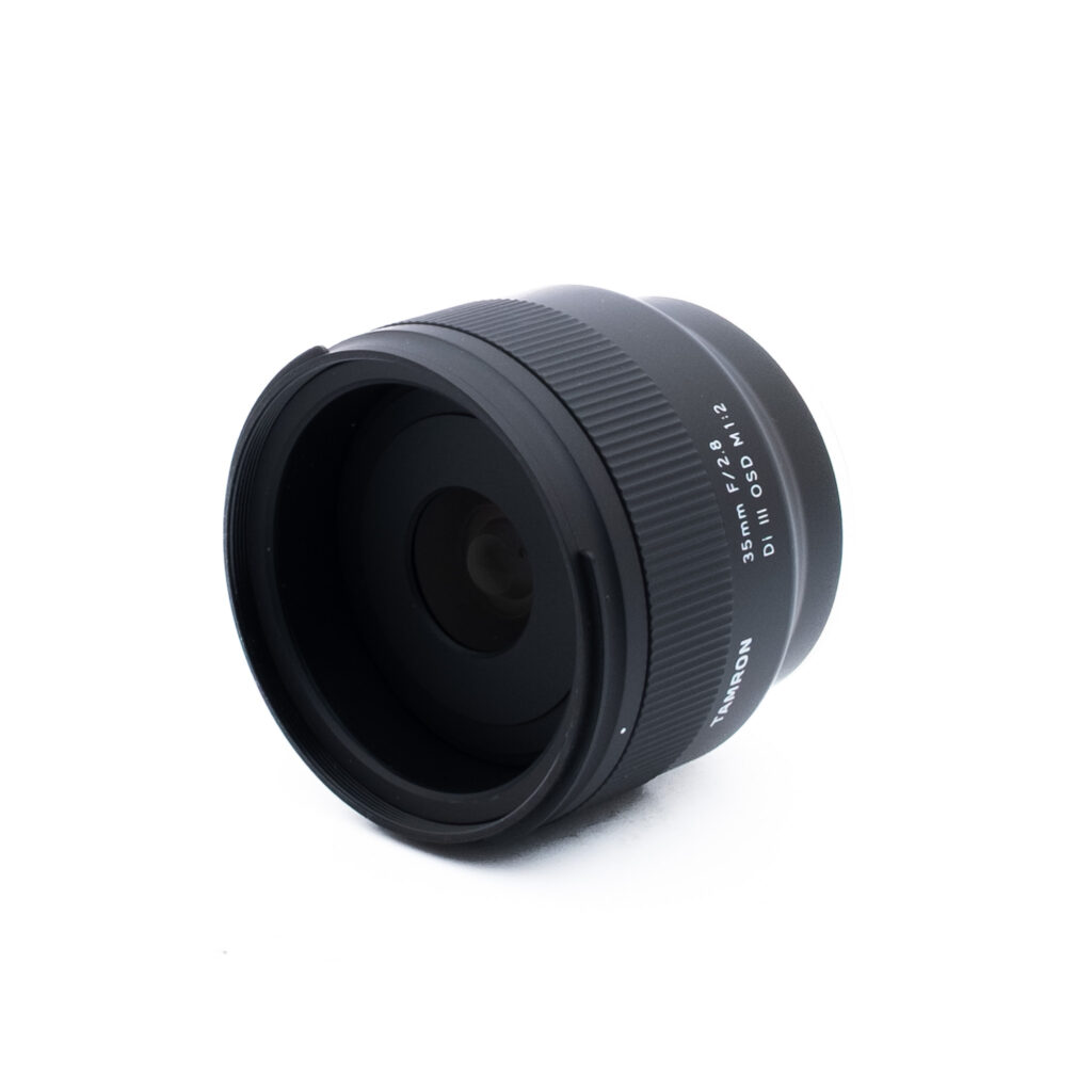 Tamron 35mm f28 Di III OSD M12 Sony-2 Musta kameran objektiivi, jossa on valkoinen teksti "35mm F2.0 W1:1.2 MC", on sijoitettu valkoiselle taustalle, ja se on taivutettu niin, että lasinen etupaneeli ja harjattu tarkennusrengas näkyvät.