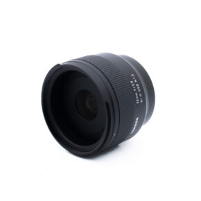 Tamron 35mm f28 Di III OSD M12 Sony-2 Musta kameran objektiivi, jossa on valkoinen teksti "35mm F2.0 W1:1.2 MC", on sijoitettu valkoiselle taustalle, ja se on taivutettu niin, että lasinen etupaneeli ja harjattu tarkennusrengas näkyvät.