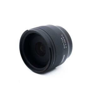 Tamron 35mm f28 Di III OSD M12 Sony-2 Musta kameran objektiivi, jossa on valkoinen teksti "35mm F2.0 W1:1.2 MC", on sijoitettu valkoiselle taustalle, ja se on taivutettu niin, että lasinen etupaneeli ja harjattu tarkennusrengas näkyvät.