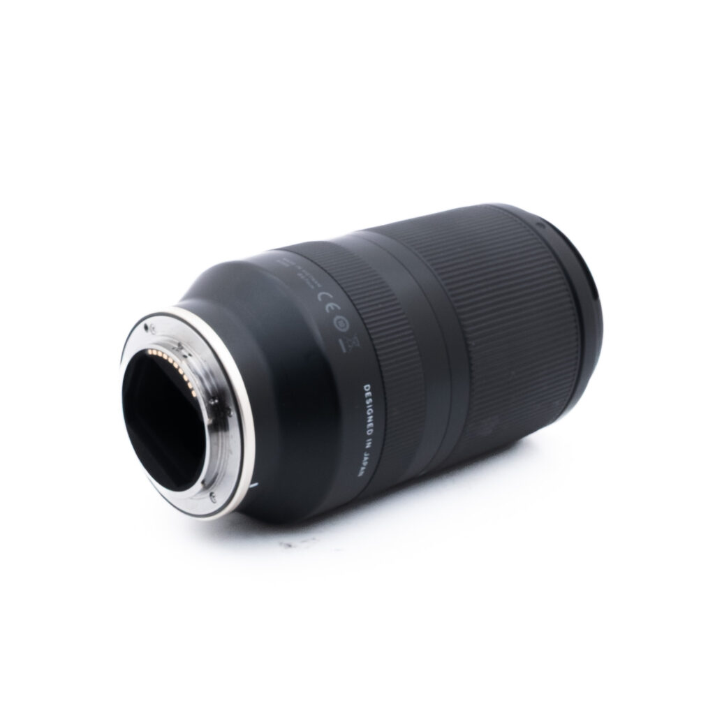Tamron 70-180mm f28 Di III VXD-1 Musta kameran linssi makaa kyljellään, vasemmassa päässä näkyvät metallinen kiinnitysrengas ja elektroniset koskettimet valkoista taustaa vasten.