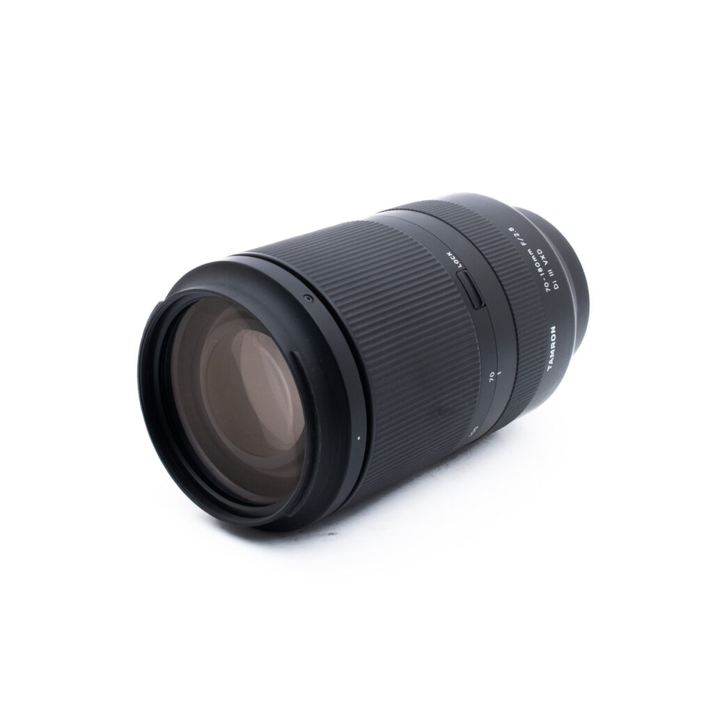 Tamron 70-180mm f28 Di III VXD-2 Musta kameran zoom-objektiivi, jossa on harjatut tarkennus- ja zoomausrenkaat, lepää valkoisella pinnalla hieman vinossa, ja siinä näkyy lasielementti ja painetut polttovälin merkinnät.