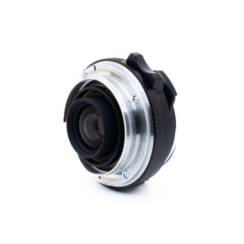 Voigtländer 35mm f25-1 Lähikuva kameran objektiivin kiinnityssovittimesta, jossa on metallisia ja mustia osia valkoisella pohjalla. Sovitin on vinossa, jolloin objektiivin aukko ja kiinnitysmekanismi näkyvät.