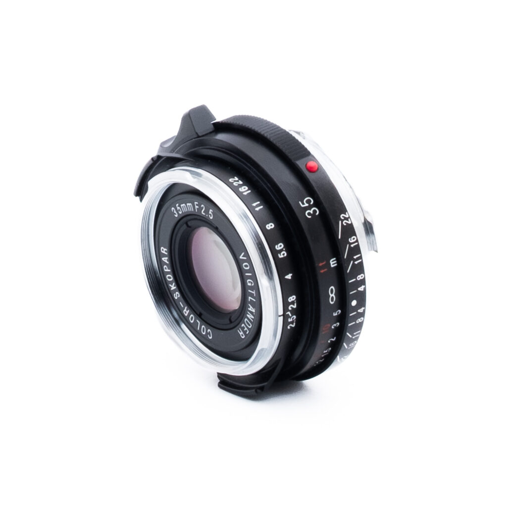 Voigtländer 35mm f25-2 Lähikuva Voigtländer Color-Skopar 35mm f/2,5 -kameran objektiivista, jossa näkyy sen lasielementti, musta ja hopeinen metallirunko sekä kaiverretut tarkennus- ja aukkomerkinnät, eristettynä valkoiselle taustalle.