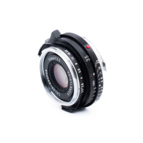 Voigtländer 35mm f25-2 Lähikuva Voigtländer Color-Skopar 35mm f/2,5 -kameran objektiivista, jossa näkyy sen lasielementti, musta ja hopeinen metallirunko sekä kaiverretut tarkennus- ja aukkomerkinnät, eristettynä valkoiselle taustalle.