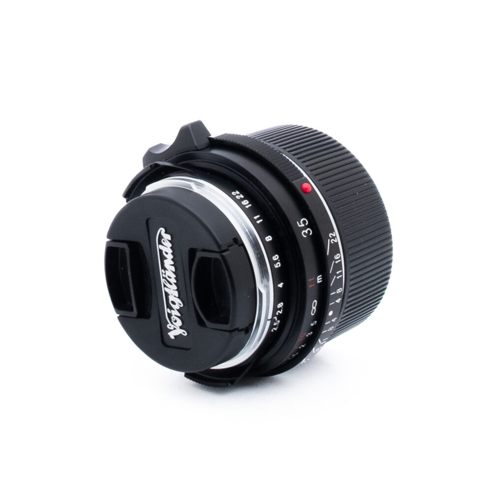 Voigtländer 35mm f25-3 Valkoisella pohjalla on Voigtländer-kameran objektiivi, jossa on musta objektiivin suojus ja jossa on merkitty aukko- ja tarkennusasetukset.