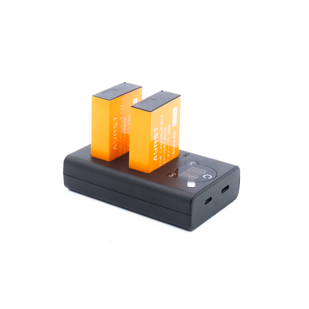 LP-E17 tarvikelaturi Cellonic Canon LP-E17 tuplalaturi, johon on asetettu kaksi oranssia Ayrst-paristoa, jossa on USB-portit ja LED-merkkivalot valkoisella pohjalla.