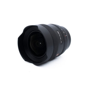 Sony 14mm f18 GM-2-3 Sony FE 14mm f/1.8 GM (Takuu 6kk) - Käytetty laajakulmaobjektiivi, jossa on suuri etuelementti ja terälehden muotoinen suojus, näkyy valkoisella taustalla.