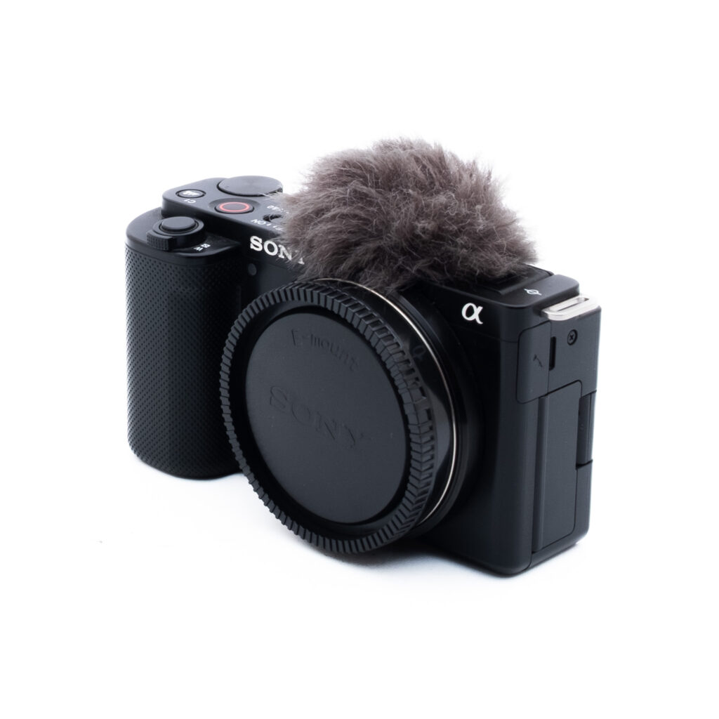 Sony ZV-E10-5 Käytetty Sony ZV-E10 (SC 51000, sis. ALV 25,5%, Takuu 6kk) kamera mustana, objektiivin suojus ja pörröinen mikrofonin tuulisuoja valkoisella pohjalla.