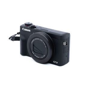 Canon G7X III-5 Käytetty Canon PowerShot G7 X Mark III (Kunto K5, Takuu 12kk) -digitaalikamera on kuvassa mustana objektiivin suojuksen ja rannehihnan kanssa valkoisella pohjalla. Kompaktikamerassa on useita ohjauspainikkeita.
