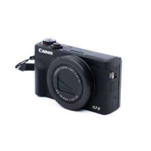 Canon G7X III-5 Käytetty Canon PowerShot G7 X Mark III (Kunto K5, Takuu 12kk) -digitaalikamera on kuvassa mustana objektiivin suojuksen ja rannehihnan kanssa valkoisella pohjalla. Kompaktikamerassa on useita ohjauspainikkeita.